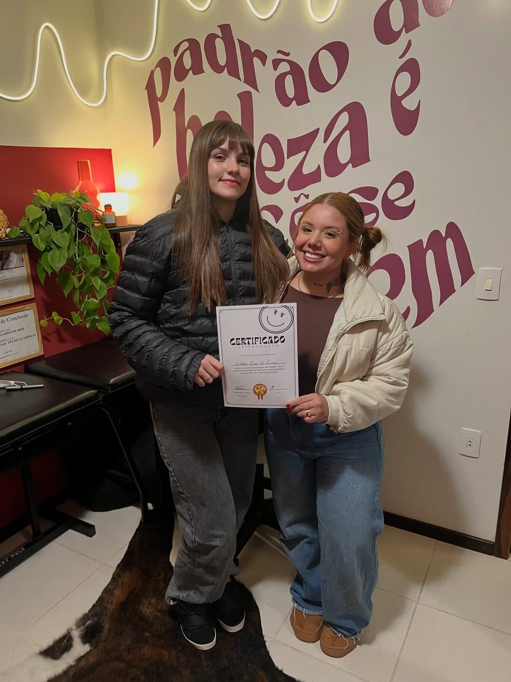 Duas alunas com o certificado do curso, em frente ao mural do estúdio